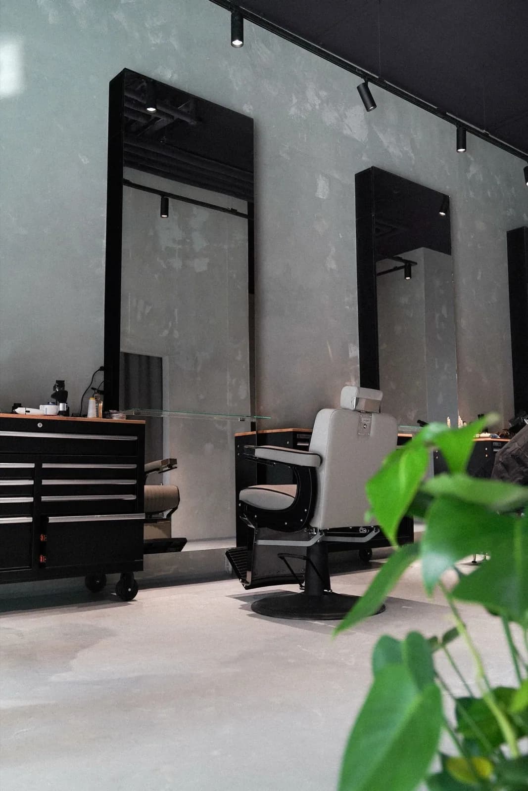 Arbeitsplatz im ONYX Cuts Salon: Barberstuhl vor schwarz gerahmtem Spiegel, Werkzeugwagen und Betonwand.